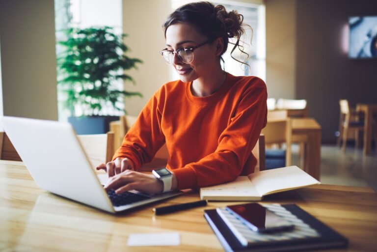 Woman in orange sweater typing on a laptop at a desk with notes and a tablet.